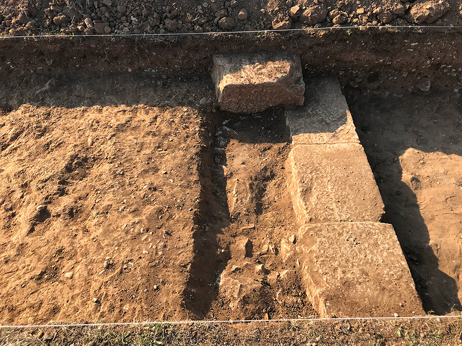 A close-up view of an archaeological excavation on the decumanus maximus. Several stone blocks are visible, including a column base lying on the southern border of the road, indicating the presence of a monumental infrastructure.