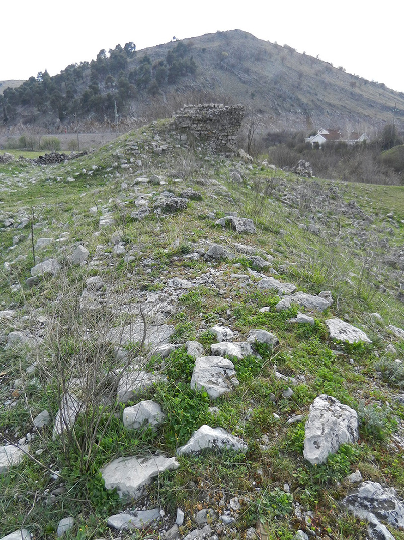 Ground view of the remains of a fortification wall in the western part of Doclea, with the ruins of a tower visible in the background. The state of preservation still shows alignments of stones attesting to the defensive structure.