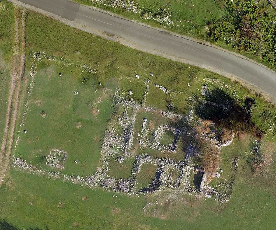 This image is an aerial orthophoto taken by a drone, showing the remains of an ancient private house. The foundations of the walls are clearly visible, forming multiple rooms organized around a central courtyard. The stone structures are partially covered by vegetation, and a modern road runs close to the archaeological site. The alignment of the walls suggests a regular architectural plan, likely influenced by Roman urban principles.