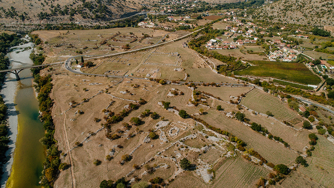 This panoramic drone view captures the entire archaeological site of Doclea, an ancient Roman city. Several ruined structures are visible, aligned according to an organized urban plan. The river bordering the site highlights the natural surroundings, while modern houses in the background illustrate the continuity of human habitation in the region.