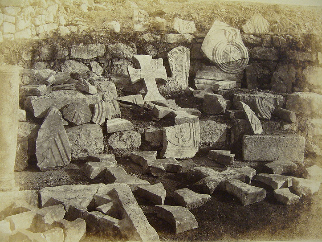 This historical photograph displays a collection of carved stone fragments found in the altar space of Basilica A. Among the remains, Christian crosses and circular decorative motifs are visible, along with architectural elements adorned with reliefs. The stones are stacked against a rough stone wall.