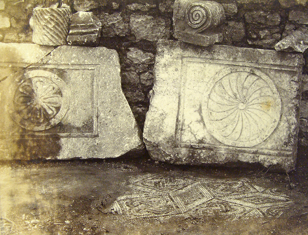 A photograph showing two large carved slabs with rosette motifs, likely part of the basilica’s decoration. On the ground, a well-preserved section of a mosaic with geometric patterns is visible. Fragments of columns and capitals can be seen in the background.