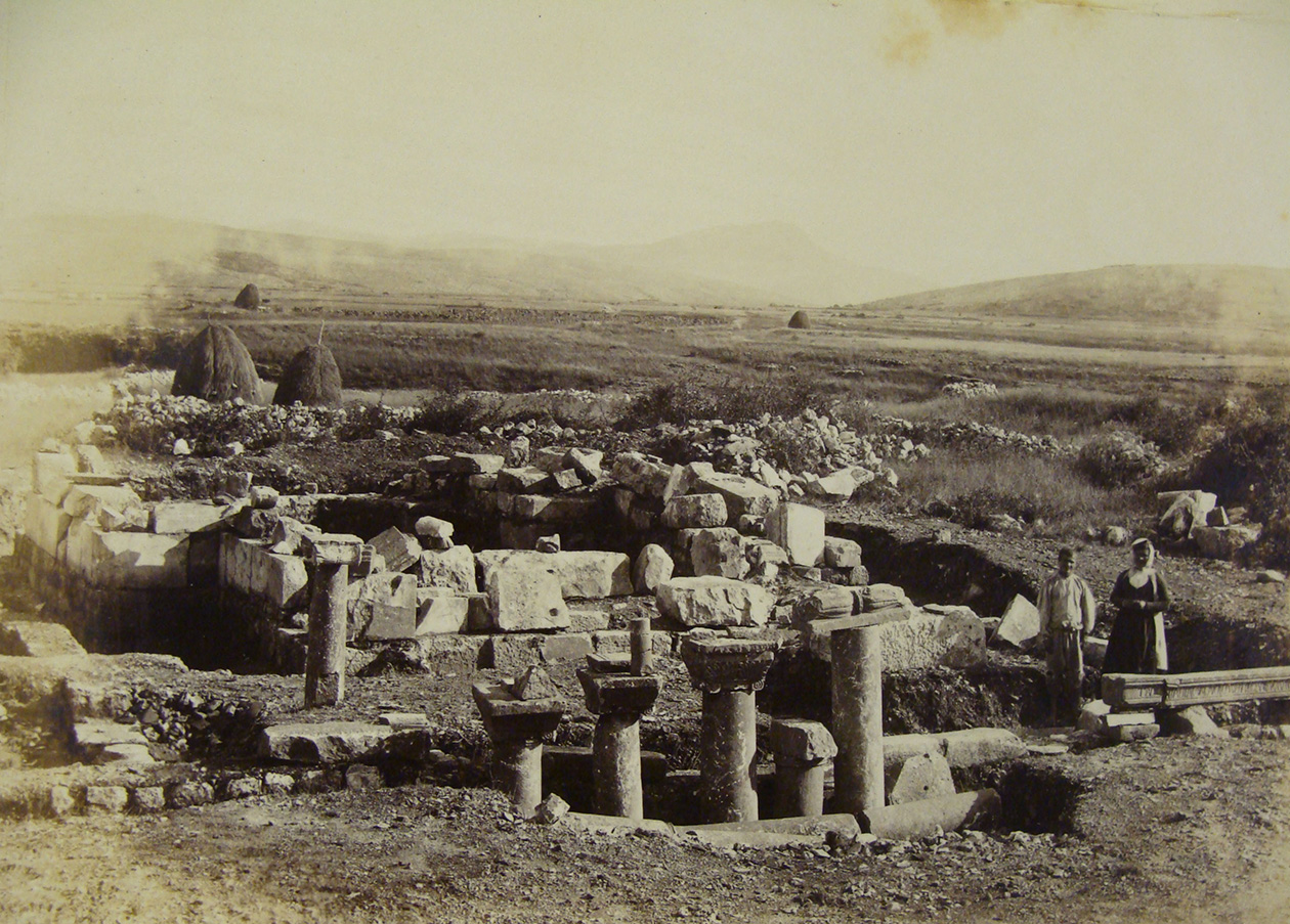 A historical photograph capturing Basilica B and the Cruciform Church from the west. Fallen columns and carved blocks are scattered in the foreground. In the background, workers or archaeologists are visible, suggesting ongoing excavations. The surrounding landscape consists of vast open plains.