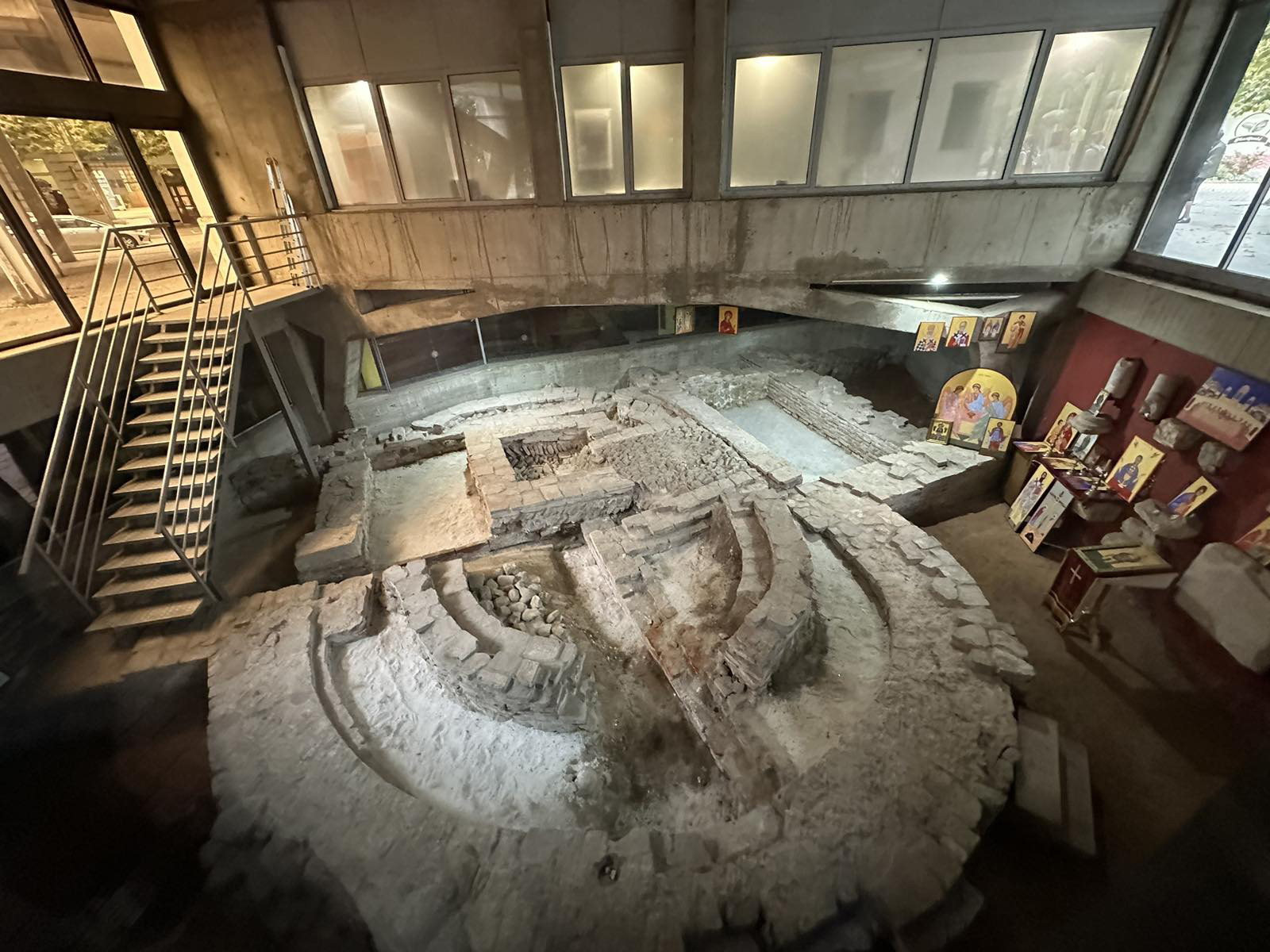 Photographie d’un site archéologique couvert présentant les vestiges d’un édifice paléochrétien, avec murs en maçonnerie conservés à faible hauteur, structures absidiales et niveaux de sol visibles, intégrés dans un bâtiment moderne