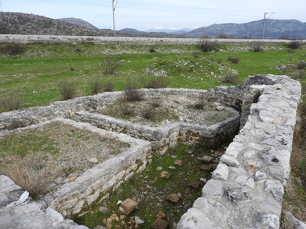 This photograph displays a stone structure with walls still preserved up to a certain height. On the ground, elements of the hypocaust system are visible, an underfloor heating method used in Roman baths and some luxurious houses. The remains of bricks and stones reveal the ingenuity of ancient construction techniques.