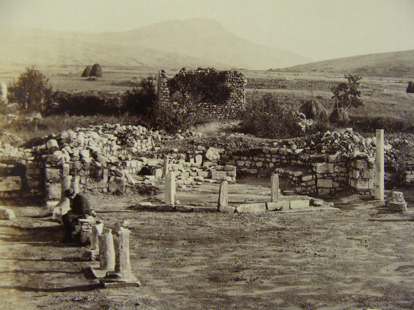 A historical photograph from 1893 showing the eastern part of Basilica A during excavation. Several columns and pillar bases are visible, some standing while others have partially collapsed. The background reveals a hilly landscape with haystacks scattered across the plain. Stone walls, partially covered by vegetation, enclose the archaeological area.