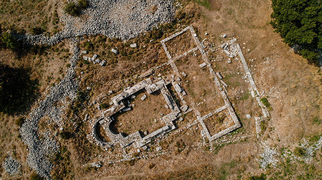 An aerial view of Basilica B and the Cruciform Church, showing well-preserved foundations. The Cruciform Church features a characteristic semicircular apse, while Basilica B follows a classic rectangular plan. The ruins are spread out in a natural environment dominated by stones and low vegetation.