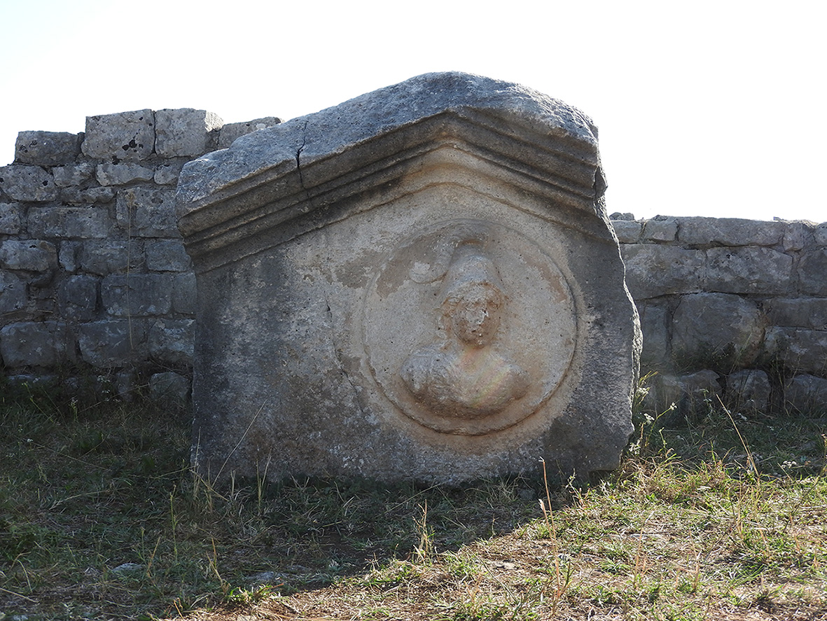 Sculpted detail of a small pediment depicting a helmeted female figure, interpreted as the goddess Roma. This relief testifies to the presence of imperial iconographic references in Doclea.