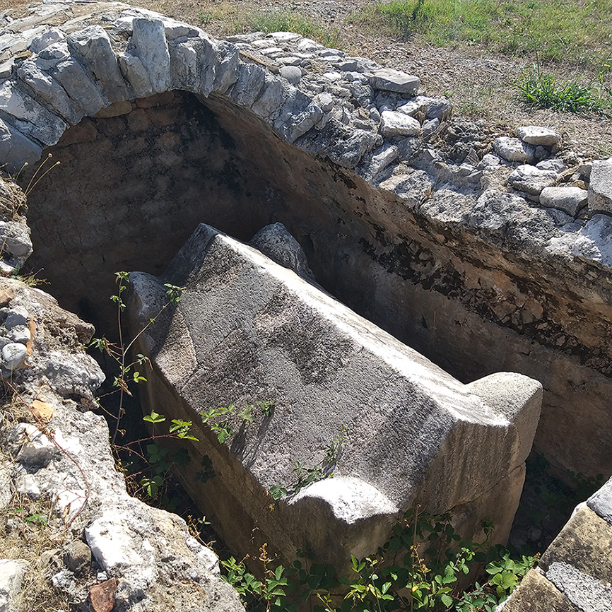 An aerial photograph of the Bjelovine archaeological site, showing the foundations of several ancient structures. Stone walls form a grid indicating urban organization. The site is surrounded by fields and sparse vegetation.